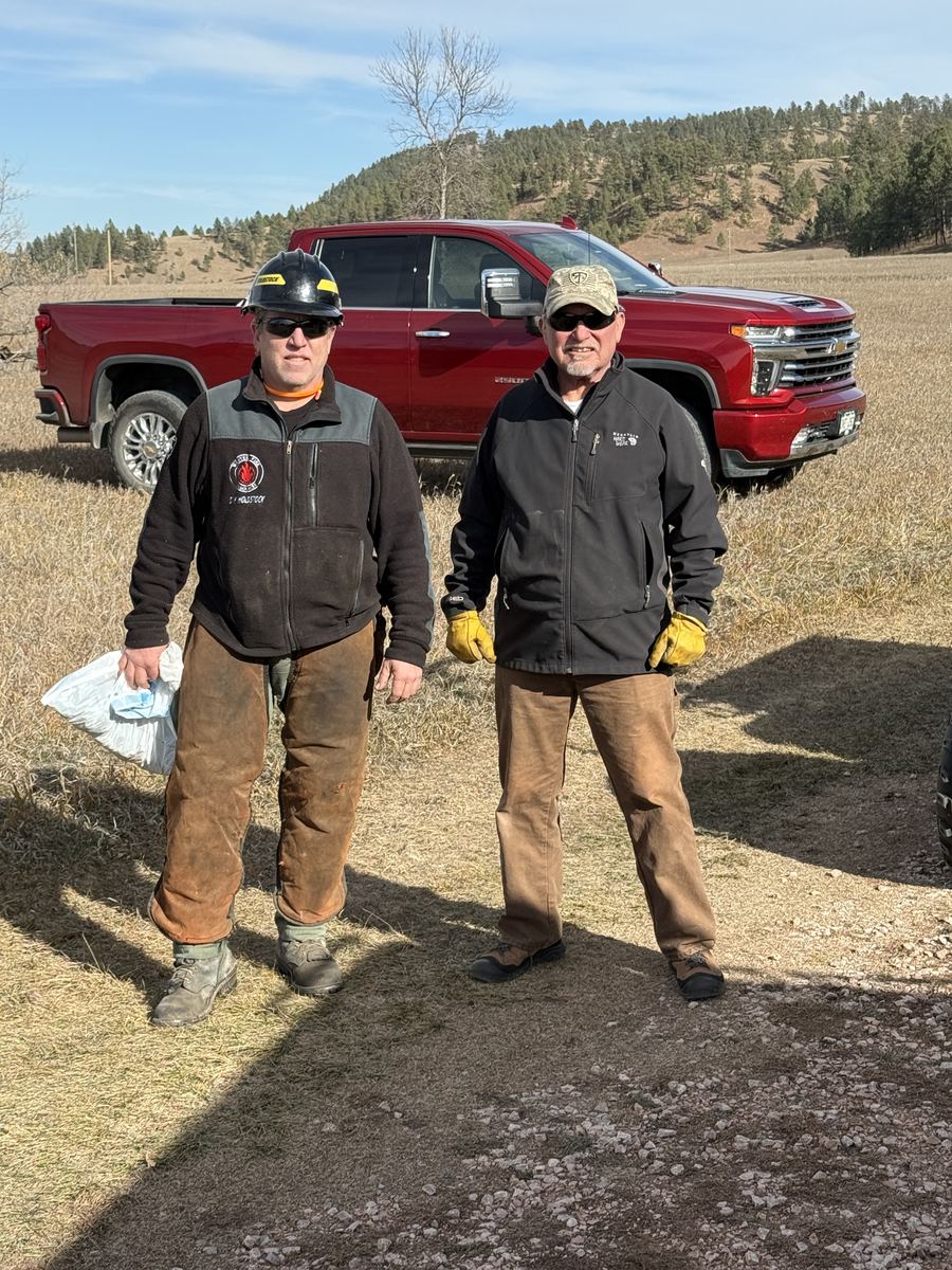 Two Wildfire Pros foresters on a project site &mdash; one in a wildland fire helmet and brush chaps, one in a cap and work gloves &mdash; standing in front of a pickup on a grassy meadow below a ponderosa pine ridge